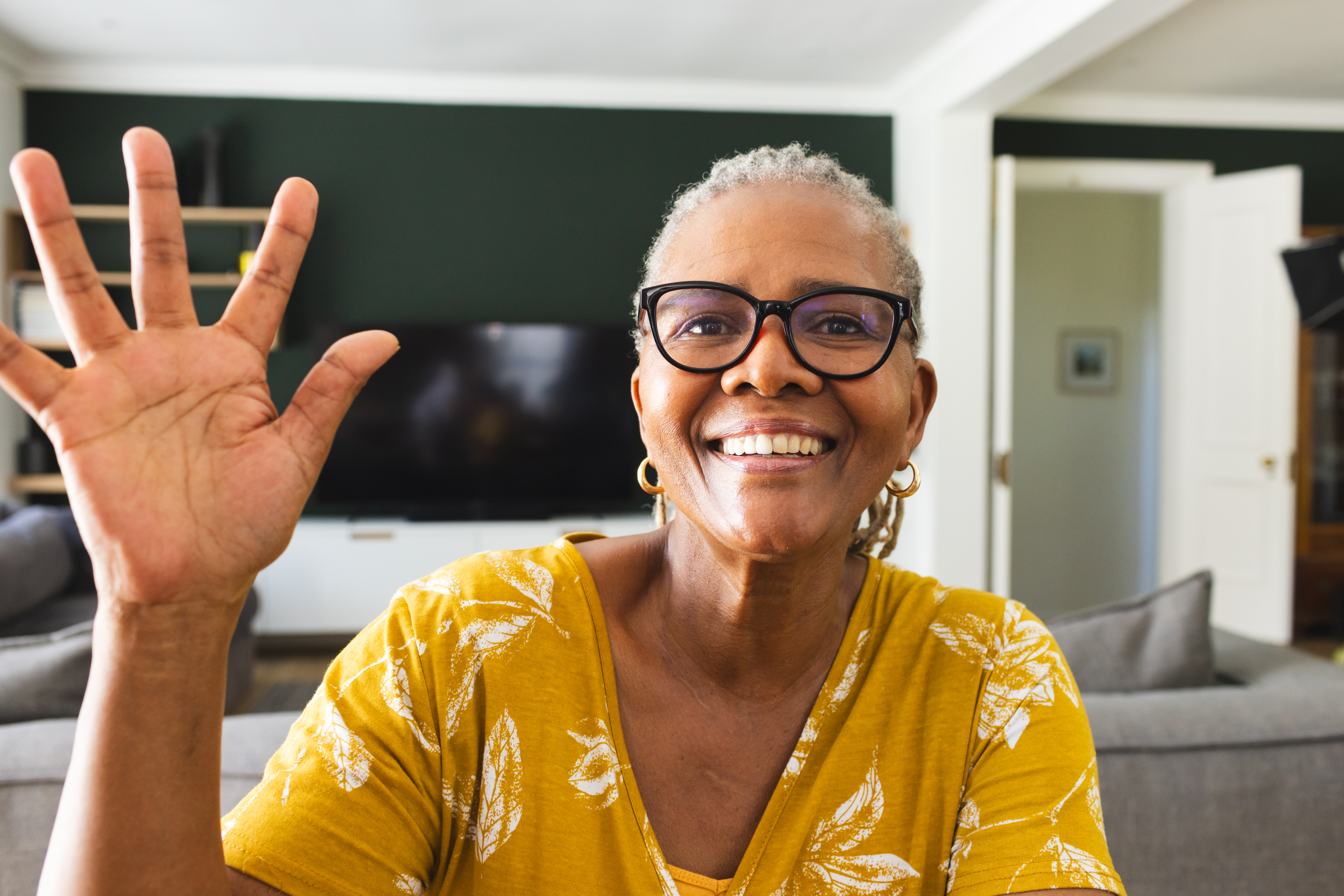 Senior African American woman waves with a bright smile, wearing glasses and a yellow top. Her joyful expression fills the cozy living room setting with warmth.
